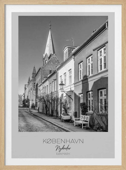 A black and white framed travel poster of a charming cobblestone street in Nyboder, Copenhagen. The photograph shows a sunlit row of historic houses with a prominent church steeple in the background. At the bottom of the poster, the words 'KØBENHAVN', 'Nyboder', and 'DANMARK' are written. Wall Art