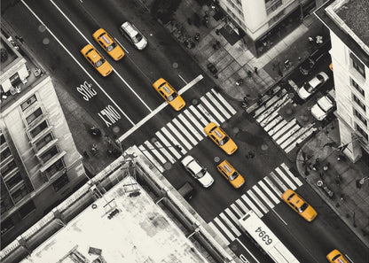 An aerial, black and white photograph of a bustling New York City intersection, with the iconic yellow taxi cabs highlighted in a vibrant selective color. Pedestrians cross the street as cars and a bus navigate the busy roads between tall buildings. Wall Art