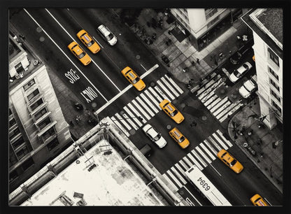 An aerial, black and white photograph of a bustling New York City intersection, with the iconic yellow taxi cabs highlighted in a vibrant selective color. Pedestrians cross the street as cars and a bus navigate the busy roads between tall buildings. Wall Art
