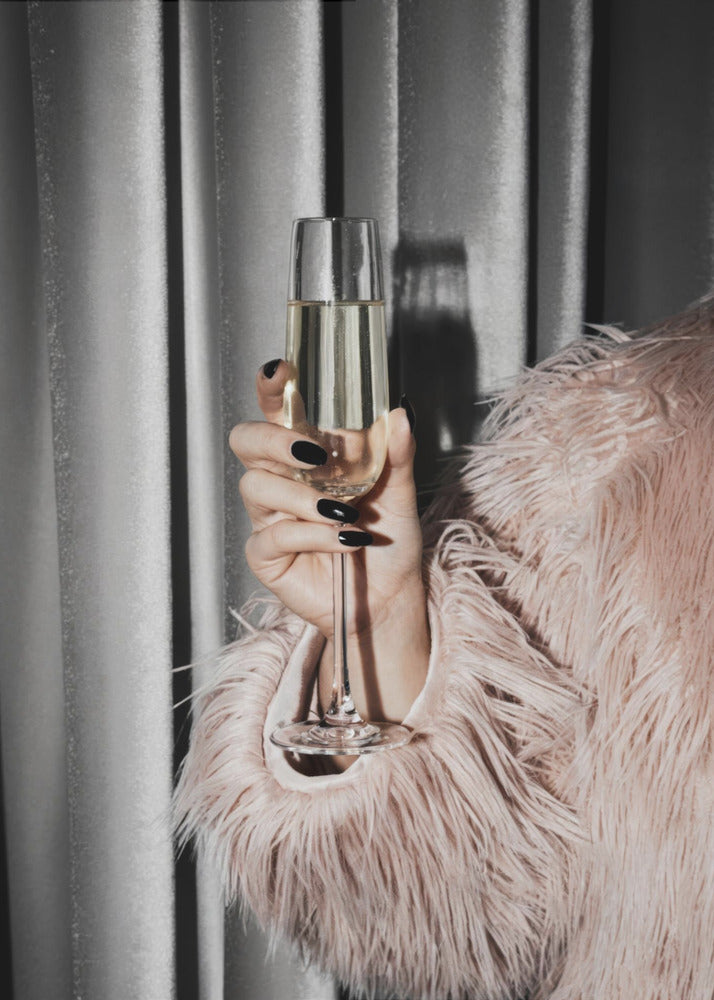 A close-up photograph of a woman's hand with black nail polish holding a tall, elegant flute of champagne. She is wearing a soft, shaggy pink faux fur coat, with a silver-gray curtain in the background, creating a luxurious and celebratory mood. Print