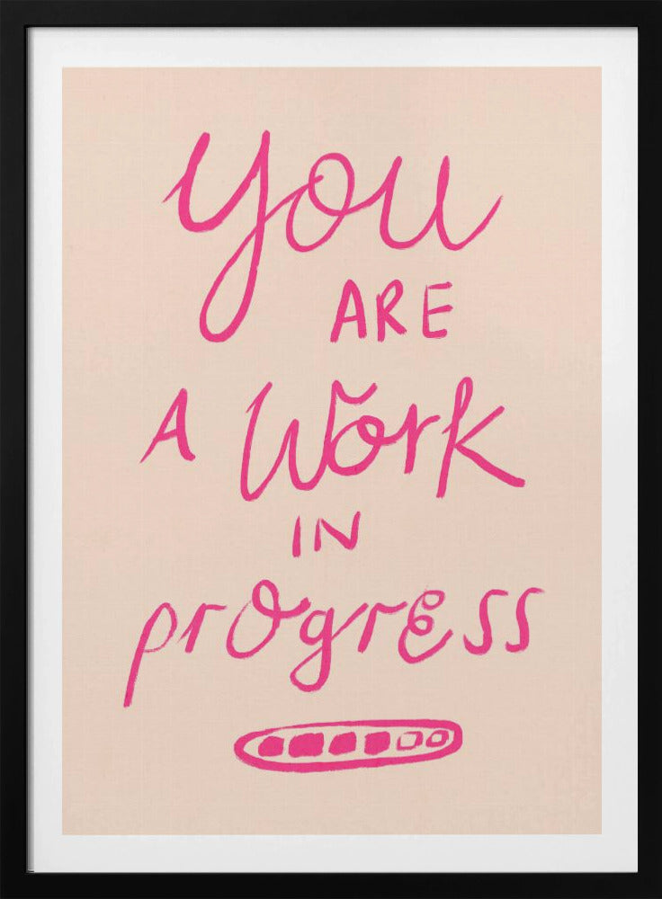 A framed art print featuring the motivational quote 'You are a work in progress'. The text is rendered in a vibrant pink, hand-lettered style against a soft beige background. The words 'You', 'a Work', and 'progress' are in a flowing script, while 'are' and 'in' are in a simple uppercase font. Wall Art