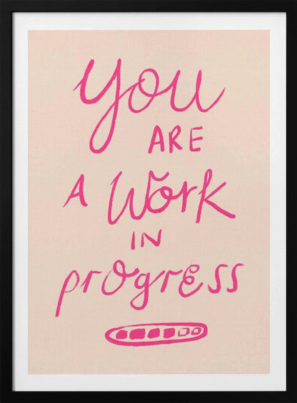 A framed art print featuring the motivational quote 'You are a work in progress'. The text is rendered in a vibrant pink, hand-lettered style against a soft beige background. The words 'You', 'a Work', and 'progress' are in a flowing script, while 'are' and 'in' are in a simple uppercase font. Wall Art