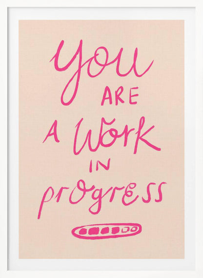 A framed art print featuring the motivational quote 'You are a work in progress'. The text is rendered in a vibrant pink, hand-lettered style against a soft beige background. The words 'You', 'a Work', and 'progress' are in a flowing script, while 'are' and 'in' are in a simple uppercase font. Wall Art