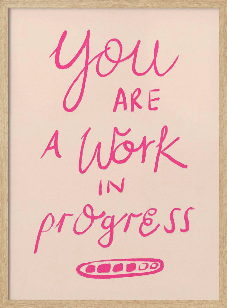 A framed art print featuring the motivational quote 'You are a work in progress'. The text is rendered in a vibrant pink, hand-lettered style against a soft beige background. The words 'You', 'a Work', and 'progress' are in a flowing script, while 'are' and 'in' are in a simple uppercase font. Wall Art