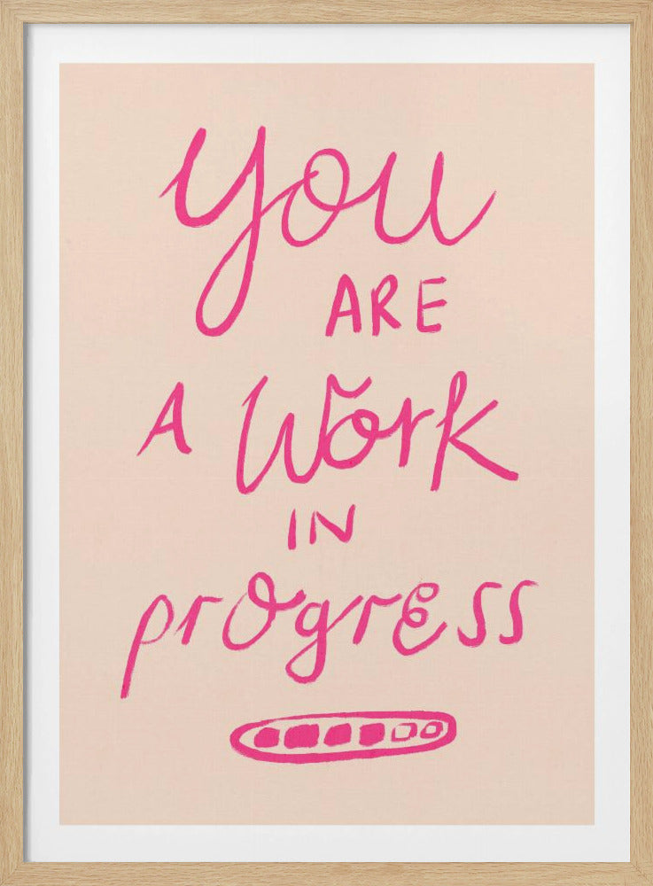 A framed art print featuring the motivational quote 'You are a work in progress'. The text is rendered in a vibrant pink, hand-lettered style against a soft beige background. The words 'You', 'a Work', and 'progress' are in a flowing script, while 'are' and 'in' are in a simple uppercase font. Wall Art