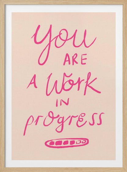 A framed art print featuring the motivational quote 'You are a work in progress'. The text is rendered in a vibrant pink, hand-lettered style against a soft beige background. The words 'You', 'a Work', and 'progress' are in a flowing script, while 'are' and 'in' are in a simple uppercase font. Wall Art