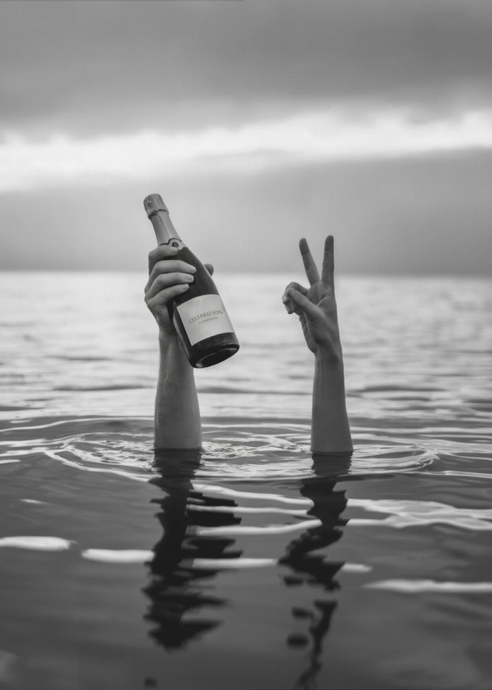 A black and white vertical photograph showing two arms sticking out of calm water. One hand holds a bottle of champagne, while the other makes a peace sign, all under a cloudy sky. Poster