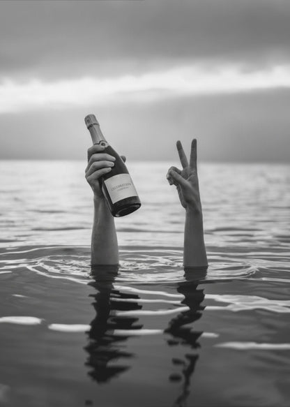 A black and white vertical photograph showing two arms sticking out of calm water. One hand holds a bottle of champagne, while the other makes a peace sign, all under a cloudy sky. Poster