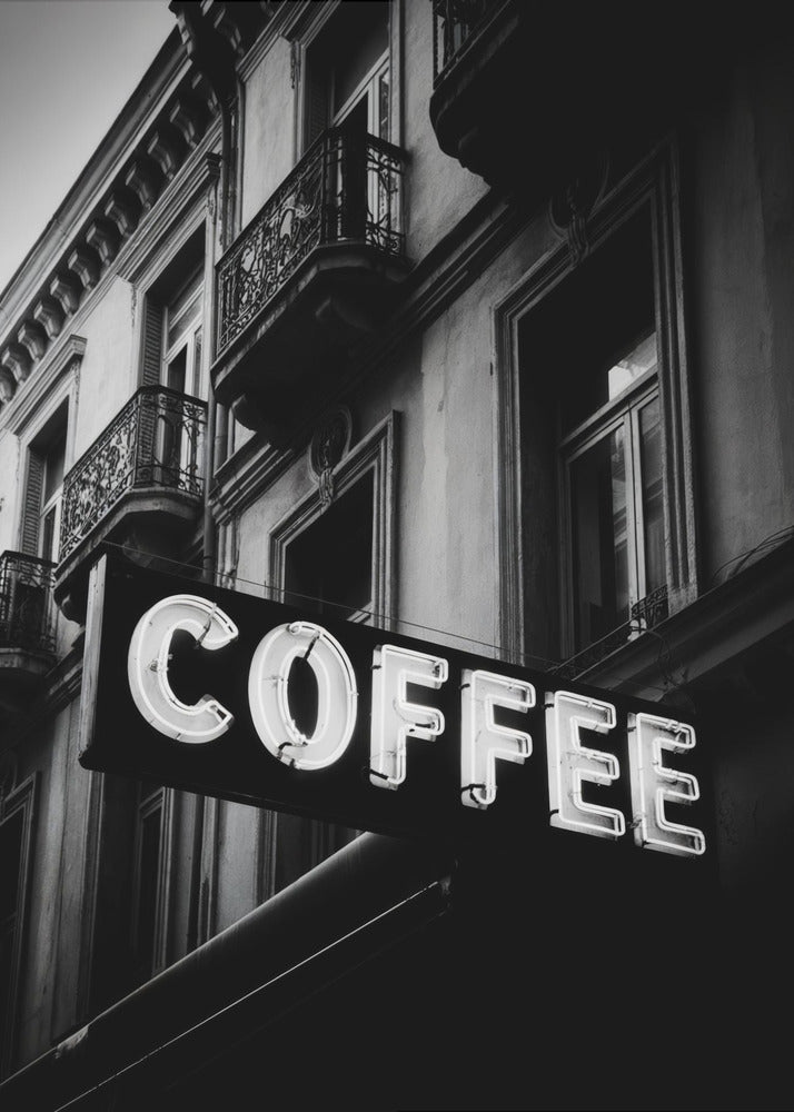 A dramatic, low-angle, black and white photograph of a bright white neon sign that reads 'COFFEE', mounted on a classic-style building with ornate balconies. Print