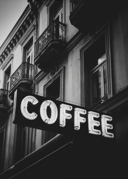 A dramatic, low-angle, black and white photograph of a bright white neon sign that reads 'COFFEE', mounted on a classic-style building with ornate balconies. Print