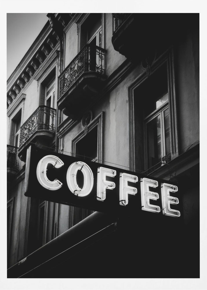 A dramatic, low-angle, black and white photograph of a bright white neon sign that reads 'COFFEE', mounted on a classic-style building with ornate balconies. Print