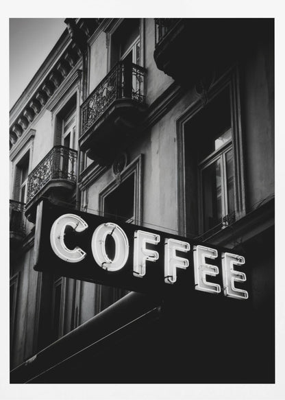 A dramatic, low-angle, black and white photograph of a bright white neon sign that reads 'COFFEE', mounted on a classic-style building with ornate balconies. Print
