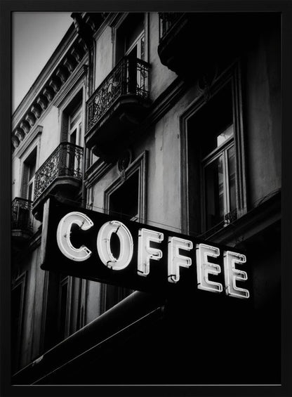 A dramatic, low-angle, black and white photograph of a bright white neon sign that reads 'COFFEE', mounted on a classic-style building with ornate balconies. Print