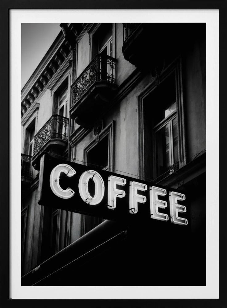 A dramatic, low-angle, black and white photograph of a bright white neon sign that reads 'COFFEE', mounted on a classic-style building with ornate balconies. Print