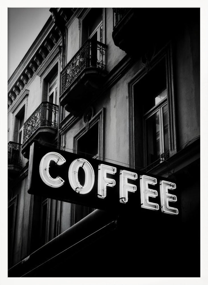 A dramatic, low-angle, black and white photograph of a bright white neon sign that reads 'COFFEE', mounted on a classic-style building with ornate balconies. Print