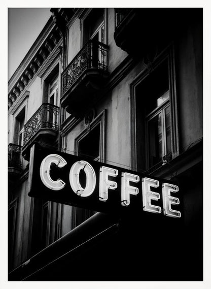 A dramatic, low-angle, black and white photograph of a bright white neon sign that reads 'COFFEE', mounted on a classic-style building with ornate balconies. Print