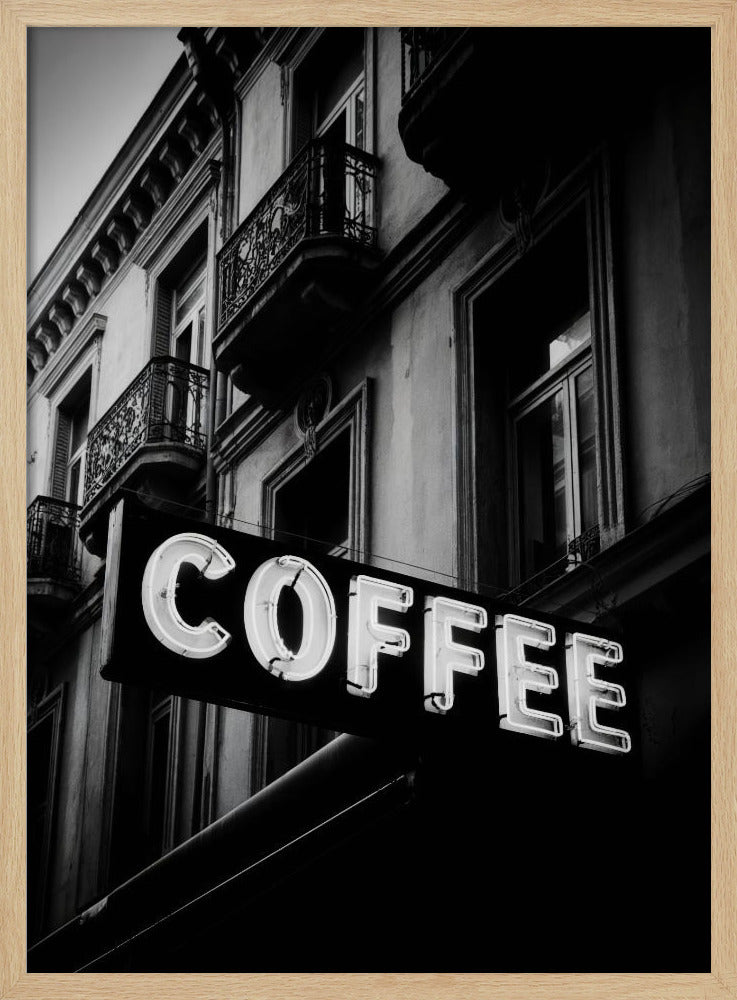 A dramatic, low-angle, black and white photograph of a bright white neon sign that reads 'COFFEE', mounted on a classic-style building with ornate balconies. Print