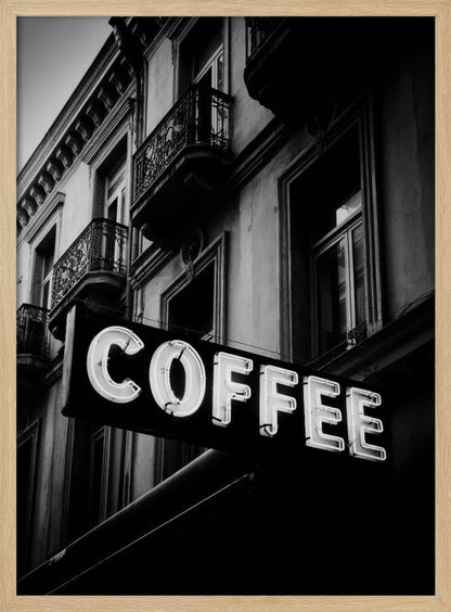A dramatic, low-angle, black and white photograph of a bright white neon sign that reads 'COFFEE', mounted on a classic-style building with ornate balconies. Print
