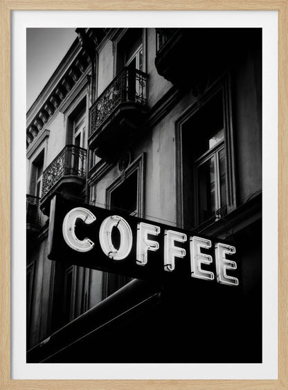 A dramatic, low-angle, black and white photograph of a bright white neon sign that reads 'COFFEE', mounted on a classic-style building with ornate balconies. Print