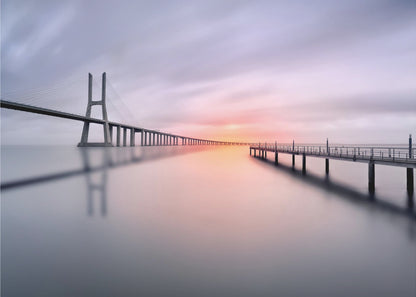 A serene long-exposure photograph of a modern cable-stayed bridge and a pier stretching over mirror-like water at sunset. The sky is filled with soft, streaky clouds in hues of pink and purple, reflected on the calm water's surface, all enclosed in a silver frame. Decor