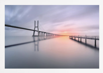 A serene long-exposure photograph of a modern cable-stayed bridge and a pier stretching over mirror-like water at sunset. The sky is filled with soft, streaky clouds in hues of pink and purple, reflected on the calm water's surface, all enclosed in a silver frame. Decor