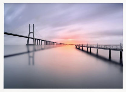 A serene long-exposure photograph of a modern cable-stayed bridge and a pier stretching over mirror-like water at sunset. The sky is filled with soft, streaky clouds in hues of pink and purple, reflected on the calm water's surface, all enclosed in a silver frame. Decor