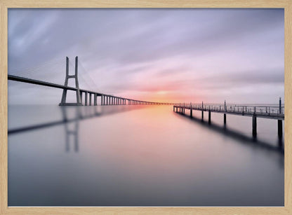 A serene long-exposure photograph of a modern cable-stayed bridge and a pier stretching over mirror-like water at sunset. The sky is filled with soft, streaky clouds in hues of pink and purple, reflected on the calm water's surface, all enclosed in a silver frame. Decor