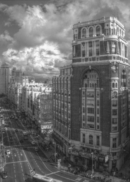 A dramatic black and white high-angle photograph of a bustling city street. A large, ornate historical building dominates the right side, while traffic flows along the avenue on the left under a sky filled with heavy, textured clouds. Poster
