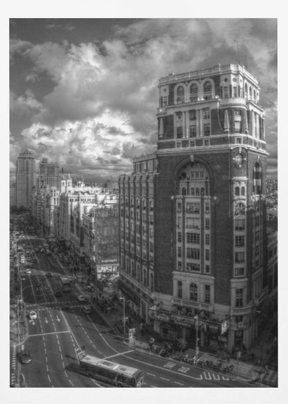 A dramatic black and white high-angle photograph of a bustling city street. A large, ornate historical building dominates the right side, while traffic flows along the avenue on the left under a sky filled with heavy, textured clouds. Poster