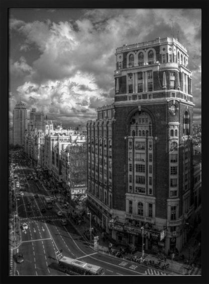 A dramatic black and white high-angle photograph of a bustling city street. A large, ornate historical building dominates the right side, while traffic flows along the avenue on the left under a sky filled with heavy, textured clouds. Poster