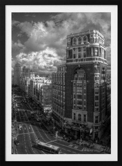 A dramatic black and white high-angle photograph of a bustling city street. A large, ornate historical building dominates the right side, while traffic flows along the avenue on the left under a sky filled with heavy, textured clouds. Poster