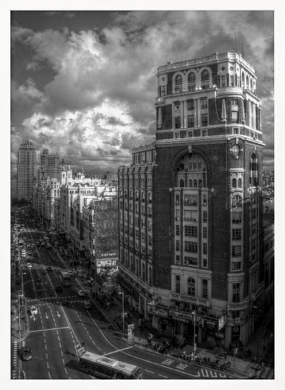 A dramatic black and white high-angle photograph of a bustling city street. A large, ornate historical building dominates the right side, while traffic flows along the avenue on the left under a sky filled with heavy, textured clouds. Poster