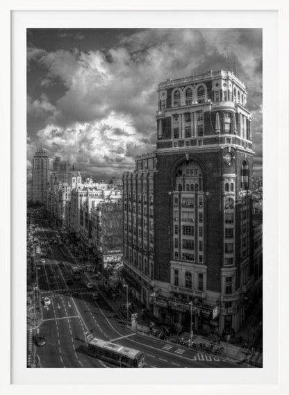 A dramatic black and white high-angle photograph of a bustling city street. A large, ornate historical building dominates the right side, while traffic flows along the avenue on the left under a sky filled with heavy, textured clouds. Poster