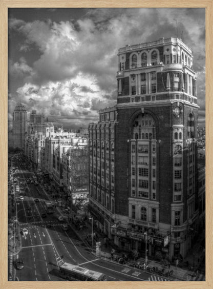 A dramatic black and white high-angle photograph of a bustling city street. A large, ornate historical building dominates the right side, while traffic flows along the avenue on the left under a sky filled with heavy, textured clouds. Poster