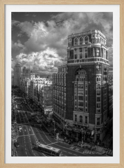 A dramatic black and white high-angle photograph of a bustling city street. A large, ornate historical building dominates the right side, while traffic flows along the avenue on the left under a sky filled with heavy, textured clouds. Poster