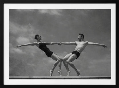 A black and white photograph of a male and female ballet dancer striking a symmetrical, athletic pose on a ledge. They hold hands, leaning away from each other in a display of strength and balance against a cloudy sky, all enclosed within a silver frame. Print