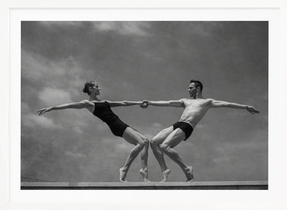 A black and white photograph of a male and female ballet dancer striking a symmetrical, athletic pose on a ledge. They hold hands, leaning away from each other in a display of strength and balance against a cloudy sky, all enclosed within a silver frame. Print