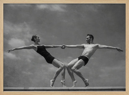 A black and white photograph of a male and female ballet dancer striking a symmetrical, athletic pose on a ledge. They hold hands, leaning away from each other in a display of strength and balance against a cloudy sky, all enclosed within a silver frame. Print