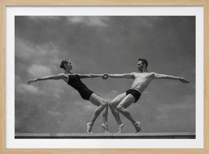 A black and white photograph of a male and female ballet dancer striking a symmetrical, athletic pose on a ledge. They hold hands, leaning away from each other in a display of strength and balance against a cloudy sky, all enclosed within a silver frame. Print