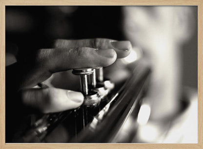 A dramatic, close-up, black and white photograph of a musician's fingers pressing down on the silver valves of a trumpet, with a soft, out-of-focus background. The image is enclosed in a brushed silver frame. Poster