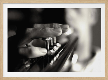 A dramatic, close-up, black and white photograph of a musician's fingers pressing down on the silver valves of a trumpet, with a soft, out-of-focus background. The image is enclosed in a brushed silver frame. Poster