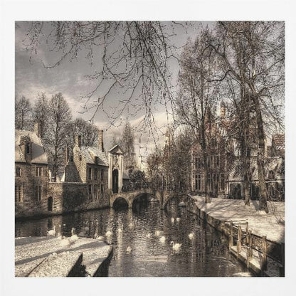 A vintage-style, framed photograph of a European canal scene in winter. Historic buildings and bare trees line the partially frozen canal, where several white swans are swimming. A light dusting of snow covers the canal banks and rooftops under a cloudy sky. Wall Art