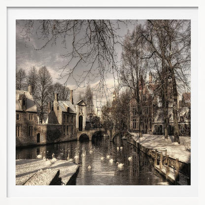 A vintage-style, framed photograph of a European canal scene in winter. Historic buildings and bare trees line the partially frozen canal, where several white swans are swimming. A light dusting of snow covers the canal banks and rooftops under a cloudy sky. Wall Art