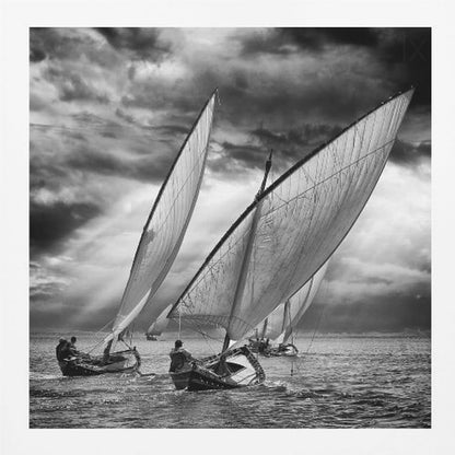 A dramatic black and white photograph of a fleet of traditional wooden sailboats with large, billowing sails racing on a choppy sea under a dark, stormy sky. The image is presented in a light wood frame. Print