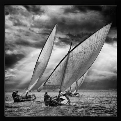A dramatic black and white photograph of a fleet of traditional wooden sailboats with large, billowing sails racing on a choppy sea under a dark, stormy sky. The image is presented in a light wood frame. Print
