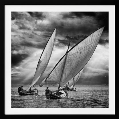 A dramatic black and white photograph of a fleet of traditional wooden sailboats with large, billowing sails racing on a choppy sea under a dark, stormy sky. The image is presented in a light wood frame. Print