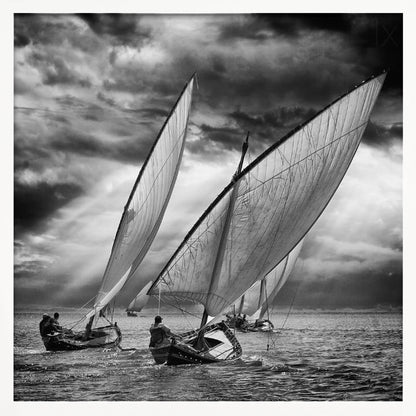 A dramatic black and white photograph of a fleet of traditional wooden sailboats with large, billowing sails racing on a choppy sea under a dark, stormy sky. The image is presented in a light wood frame. Print