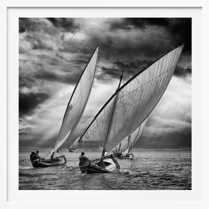 A dramatic black and white photograph of a fleet of traditional wooden sailboats with large, billowing sails racing on a choppy sea under a dark, stormy sky. The image is presented in a light wood frame. Print