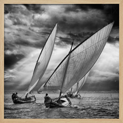 A dramatic black and white photograph of a fleet of traditional wooden sailboats with large, billowing sails racing on a choppy sea under a dark, stormy sky. The image is presented in a light wood frame. Print