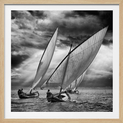 A dramatic black and white photograph of a fleet of traditional wooden sailboats with large, billowing sails racing on a choppy sea under a dark, stormy sky. The image is presented in a light wood frame. Print